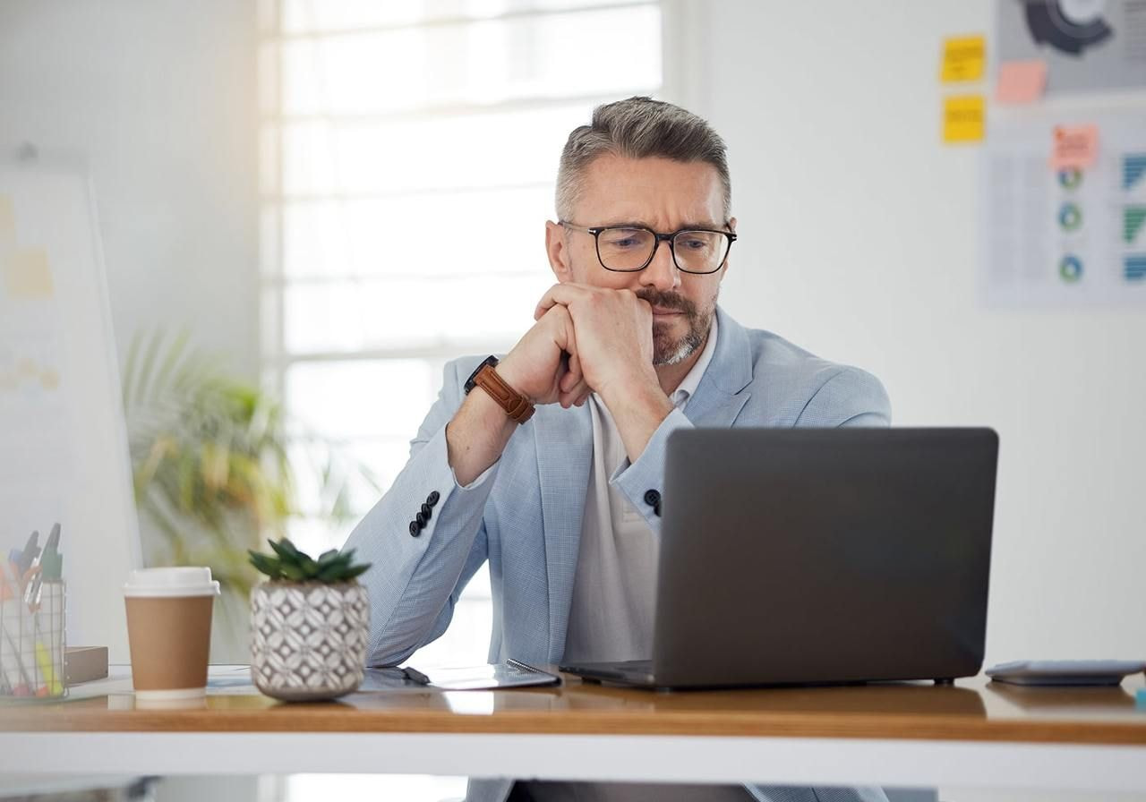 A man wearing glasses is seated at a desk, working on a laptop.